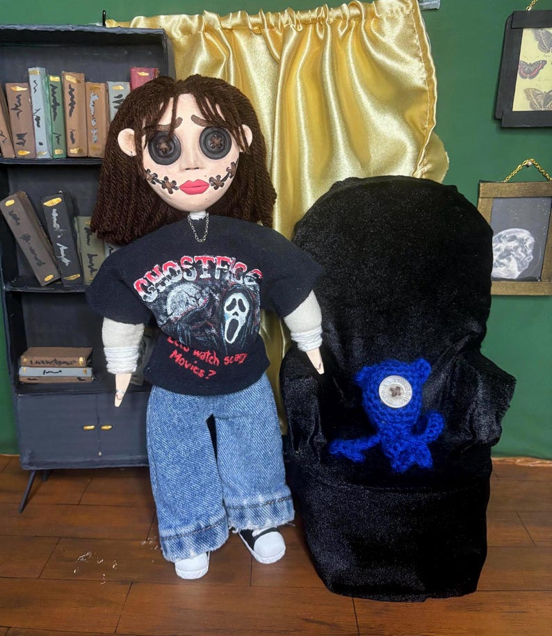 May include: A doll with button eyes, stitched mouth, and brown hair, wearing a black t-shirt with a Scream movie graphic and the text "...watch scary Movies?". The doll is wearing blue jeans and white sneakers. A blue crocheted creature sits in a black chair.