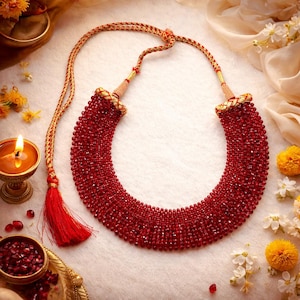 Può includere: Una vivace collana di perline rosse con nappa e dettagli dorati. La collana è composta da numerose piccole perline rotonde, creando una forma piena e curva. La nappa e il cordoncino sono rossi e dorati. La collana è esposta su una superficie bianca.