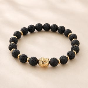 May include: Black lava stone bead bracelet with gold-colored accents. The bracelet features a gold-colored Buddha head charm. The beads are round and have a textured surface. A stylish accessory for everyday wear.