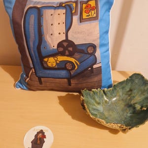 May include: A square throw pillow featuring a blue wingback chair with a sleeping yellow dog, a blue-rimmed ceramic bowl with a textured green and blue interior, and a circular sticker with the text JOANNE ILLUSTRATION.