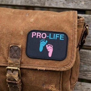 May include: Brown canvas messenger bag with a black patch that reads "PRO-LIFE" in pink and blue. The patch features blue and pink footprints. The bag has a leather strap and buckle.