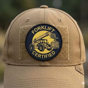 Könnte beinhalten: Eine beigefarbene Baseballkappe mit einem runden Aufnäher auf der Vorderseite. Der Aufnäher hat einen schwarzen Hintergrund mit einem gelben Kreis, einer Grafik eines Gabelstaplers, der eine Kiste hebt, und dem Text FORKLIFT CERTIFIED in Gelb.