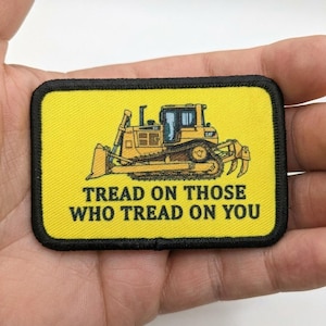 Könnte beinhalten: Ein gelber Aufnäher mit schwarzem Rand, der eine gelbe Bulldozer-Illustration zeigt. Der Text "TREAD ON THOSE WHO TREAD ON YOU" ist in Schwarz gedruckt. Der Bulldozer hat das CAT-Logo.