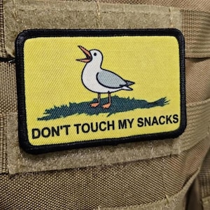 Peut inclure: Écusson rectangulaire à bordure noire sur fond jaune. Il représente une mouette de dessin animé, bec ouvert, sur de l'herbe verte. Le texte « DON'T TOUCH MY SNACKS » est en dessous.