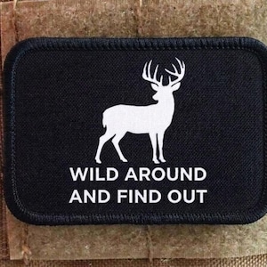 Può includere: Toppa rettangolare nera con una silhouette bianca di cervo e la scritta "WILD AROUND AND FIND OUT". La toppa è cucita su uno sfondo di tessuto beige.