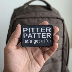 May include: A black rectangular patch with white text that reads "PITTER PATTER let's get at 'er". The patch is held in front of a brown backpack. The patch is likely an accessory for a backpack or other gear.