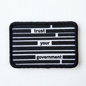 May include: Black and white rectangular patch with the words "trust your government" in a bold, sans-serif font. The patch has a black border and horizontal white stripes.
