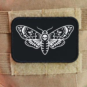Puede incluir: Un parche rectangular negro con un diseño de polilla blanca, que incluye una calavera en su cuerpo. La polilla tiene patrones de alas detallados. El parche está adherido a un fondo de tela beige.
