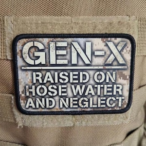 Peut inclure: Un écusson rectangulaire avec le texte "GEN-X" au-dessus de "RAISED ON HOSE WATER AND NEGLECT" en lettres blanches. L'écusson a une bordure noire et un fond camouflage, sur un tissu beige.