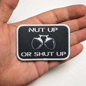 Könnte beinhalten: Schwarzer rechteckiger Aufnäher mit weißem Text und weißem Rand. Der Text lautet "NUT UP OR SHUT UP" mit einer Grafik von zwei Eicheln. Der Aufnäher wird in einer Hand gehalten.