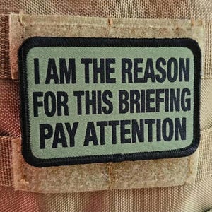 May include: A rectangular patch with the text "I AM THE REASON FOR THIS BRIEFING PAY ATTENTION" in black letters on a green background. The patch is attached to a tan fabric.