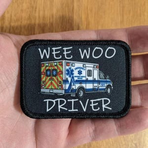 Op de afbeelding: Zwarte rechthoekige patch met een ambulance-illustratie en de tekst "WEE WOO DRIVER" in het wit. De ambulance is wit en blauw met rode en gele accenten. De patch heeft een zwarte rand.