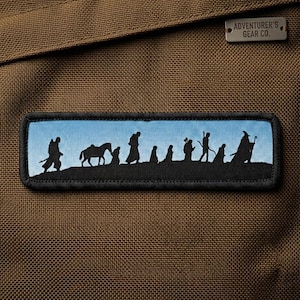 Puede incluir: Tela marrón con un parche rectangular que muestra una silueta de figuras y un caballo contra un cielo azul. El parche tiene un borde negro. Una pequeña etiqueta metálica dice "ADVENTURER'S GEAR CO."