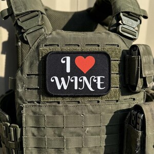 Può includere: Gilet tattico verde oliva con più tasche e cinghie. Una toppa nera con le parole "I ❤️ WINE" in bianco e un cuore rosso è attaccata alla parte anteriore del gilet.