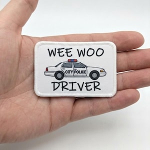 Pode incluir: Um patch retangular branco com as palavras "WEE WOO" acima de um carro de polícia de desenho animado e a palavra "DRIVER" abaixo. O carro de polícia é branco com janelas pretas e "CITY POLICE" na lateral.