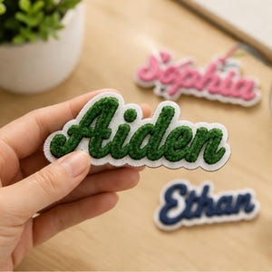 May include: A custom name patch featuring the name Aiden embroidered in dark green thread with a white border. The patch has a textured, raised stitch finish and a scalloped edge. Additional patches with the names Sophia and Ethan are visible.