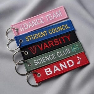 Könnte beinhalten: Fünf rechteckige bestickte Schlüsselanhänger mit silbernen Metallringen. Der rosa Anhänger zeigt DANCE TEAM, der blaue STUDENT COUNCIL, der schwarze V VARSITY, der grüne SCIENCE CLUB mit Atomsymbol und der rote BAND mit einer Musiknote.