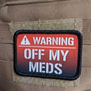 May include: A rectangular morale patch with a black border and a red-to-black gradient background. The text reads WARNING in white with an exclamation point icon, followed by the white text OFF MY MEDS. The patch is attached to tan fabric.