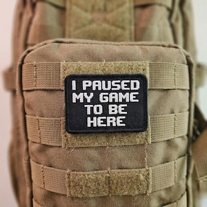 以下が含まれることがあります： 「I PAUSED MY GAME TO BE HERE」の白い文字が黒い背景に書かれた長方形のパッチ。 パッチは、複数のストラップが付いたタン色の生地に取り付けられています。