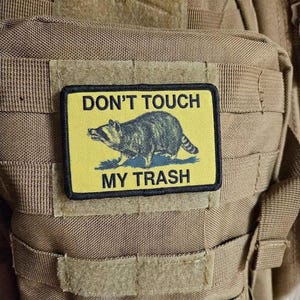 Puede incluir: Parche rectangular con el texto "DON'T TOUCH MY TRASH" y una ilustración de mapache. El parche tiene un fondo amarillo, texto negro y un borde negro. El mapache es gris y negro.