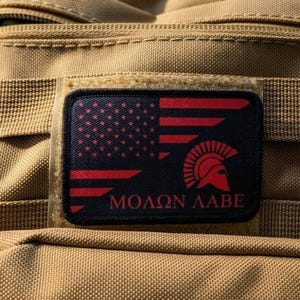 May include: A rectangular morale patch attached to a tan tactical backpack. The patch features a black background with a red American flag design, a Spartan helmet icon, and the Greek text "ΜΟΛΩΝ ΛΑΒΕ" printed in red at the bottom.