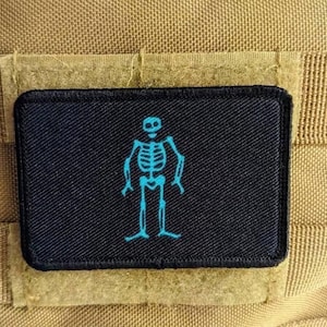 以下が含まれることがあります： 中央に水色のスケルトンの刺繍が施された長方形の黒いワッペン。ワッペンにはステッチ入りの縁取りがあり、水平なウェビングストラップが付いたベージュのテクスチャ生地の表面に取り付けられています。