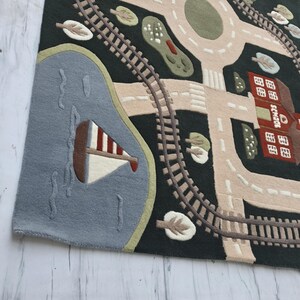 Interactive Montessori play rug featuring an illustrated town map with realistic train tracks, smooth roadways, colorful neighborhood homes, a red schoolhouse, landscaped trees, roundabout intersection, and a blue lake with a sailboat. This children's educational rug is styled inside a bright contemporary dining room, creating a beautiful combination of family-friendly design and practical learning.