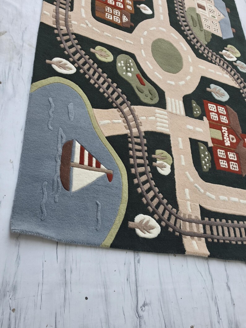 Interactive Montessori play rug featuring an illustrated town map with realistic train tracks, smooth roadways, colorful neighborhood homes, a red schoolhouse, landscaped trees, roundabout intersection, and a blue lake with a sailboat. This children's educational rug is styled inside a bright contemporary dining room, creating a beautiful combination of family-friendly design and practical learning.