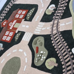 Large kids activity rug designed with an engaging transportation-themed town including curved railway tracks, interconnected roads, family homes, colorful buildings, school, landscaped greenery, pedestrian crossings, central traffic circle, and a waterfront lake with sailboat. The soft textured rug is displayed beneath a contemporary dining table, highlighting its versatility as both a functional play mat and stylish home accent.