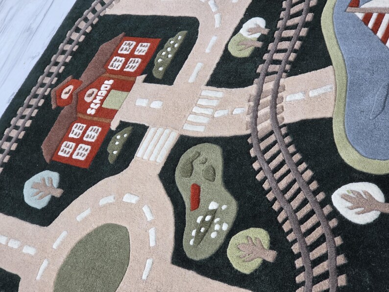Large kids activity rug designed with an engaging transportation-themed town including curved railway tracks, interconnected roads, family homes, colorful buildings, school, landscaped greenery, pedestrian crossings, central traffic circle, and a waterfront lake with sailboat. The soft textured rug is displayed beneath a contemporary dining table, highlighting its versatility as both a functional play mat and stylish home accent.