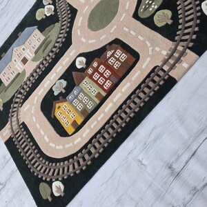 Premium educational play rug featuring a charming miniature community complete with winding roads, railway loop, neighborhood school, modern family home, colorful townhouses, decorative trees, green landscaping, and a peaceful lake with sailboat. Located beneath a modern dining table in a Scandinavian-inspired home, the rug creates an inviting family space where children can safely enjoy pretend play with trains, toy cars, trucks, and wooden vehicles.