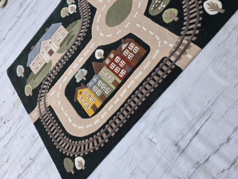 Premium educational play rug featuring a charming miniature community complete with winding roads, railway loop, neighborhood school, modern family home, colorful townhouses, decorative trees, green landscaping, and a peaceful lake with sailboat. Located beneath a modern dining table in a Scandinavian-inspired home, the rug creates an inviting family space where children can safely enjoy pretend play with trains, toy cars, trucks, and wooden vehicles.