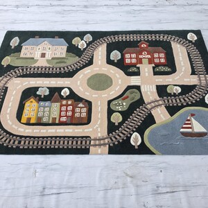 Educational Kids Road Map Rug showcasing a detailed town scene with a railway loop, school building, family house, colorful residential streets, scenic pond, sailboat, trees, and road network for endless imaginative play. This soft playmat encourages fine motor skills, creative thinking, role-playing, and early childhood development while providing a cozy play surface.