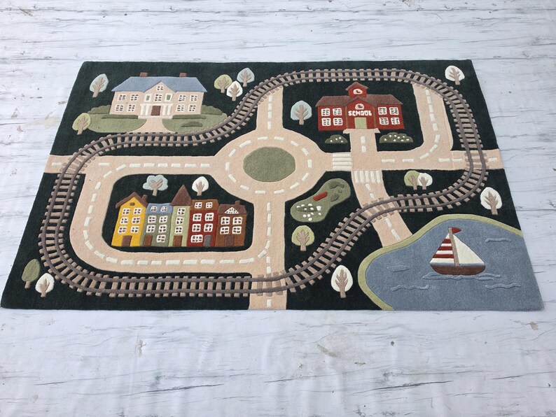 Educational Kids Road Map Rug showcasing a detailed town scene with a railway loop, school building, family house, colorful residential streets, scenic pond, sailboat, trees, and road network for endless imaginative play. This soft playmat encourages fine motor skills, creative thinking, role-playing, and early childhood development while providing a cozy play surface.