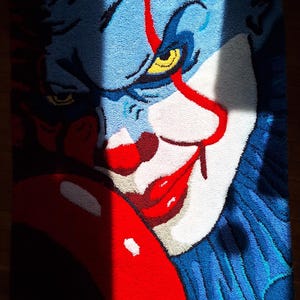 Könnte beinhalten: Blau-rot-weißer Teppich mit einem detaillierten Bild eines Clown-Gesichts. Der Teppich hat ein strukturiertes Aussehen, wobei das Gesicht des Clowns den größten Teil einnimmt. Der Clown hat rote Lippen, eine rote Nase und gelbe Augen.