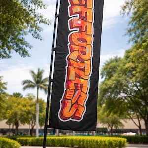 May include: A black rectangular flag with the word "EFFORT" in orange graffiti-style lettering. The flag is mounted on a black pole and stands on a grassy lawn, with trees and a blue sky in the background.