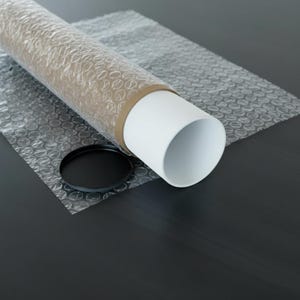 May include: A white cylindrical poster tube with a black cap, resting on bubble wrap. The tube is secured with a brown band. The scene is set on a dark surface, suggesting a focus on packaging and protection.