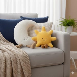 May include: A plush, cream-colored crescent moon and a yellow sun-shaped pillow with smiling faces sit on a gray sofa. The sun has eight rays and brown feet. A beige cable-knit throw and navy blue pillows complete the cozy scene.