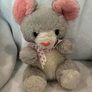 May include: A plush gray mouse toy with pink inner ears, a pink nose, and a pink and white patterned bow. The toy has black whiskers and white fur on its belly and feet.