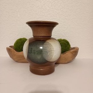May include: A brown ceramic vase with a wide, flared rim and a rounded body featuring overlapping circular patterns in shades of dark green, grey, and cream. The base is cylindrical, and the surface has a smooth, glazed finish on the midsection.