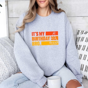 May include: A light gray sweatshirt with the text "IT'S MY BIRTHDAY BRO." in orange and yellow. The graphic includes a bicycle, a cake, and a party hat. The sweatshirt is a casual, comfortable style.