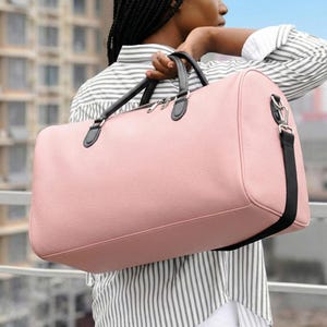 Puede incluir: Una bolsa de lona rosa con asas negras y una correa para el hombro negra. La bolsa tiene una superficie texturizada y una cremallera plateada. La bolsa se lleva sobre el hombro.