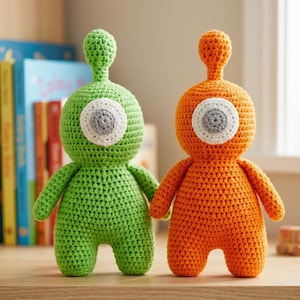 Puede incluir: Dos muñecos alienígenas de ganchillo. Uno es verde, el otro naranja. Cada uno tiene un ojo grande y único con un diseño blanco, gris y blanquecino. Tienen brazos pequeños y un cuerpo redondeado. Los muñecos están sobre una superficie de madera.