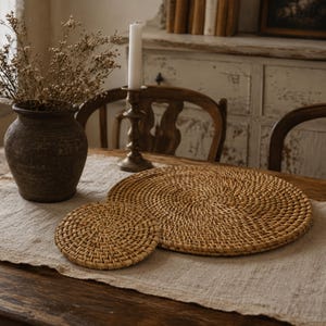 May include: Two woven rattan placemats, one large and one small, sit on a linen tablecloth. A dark brown ceramic vase with dried flowers and a brass candlestick with a white candle are also on the table. The table is made of dark wood.