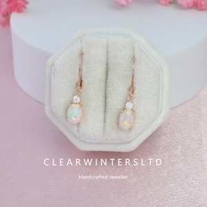 May include: Rose gold earrings with oval opal stones and a small clear stone above each opal. The earrings are displayed in a white velvet jewelry box. The text "CLEARWINTERSLTD" and "Handcrafted Jeweller" are visible.