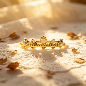 May include: A gold crown ring with a detailed design. The ring is adorned with small gemstones in clear, white, and purple hues. The ring sits on a textured surface, with scattered leaves adding to the aesthetic. A beautiful piece of jewellery.