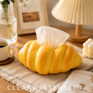 May include: A yellow tissue box cover shaped like a croissant with a textured, golden-brown crust finish. The cover features a central opening at the top where white tissues are dispensed, mimicking the appearance of a freshly baked pastry.
