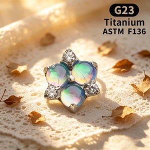 Peut inclure: Un bijou argenté en forme de fleur avec trois gemmes sphériques irisées et des pierres d'accent claires. Le texte "G23 Titanium ASTM F136" est visible. Posé sur une surface en dentelle blanche avec des feuilles sèches éparpillées.