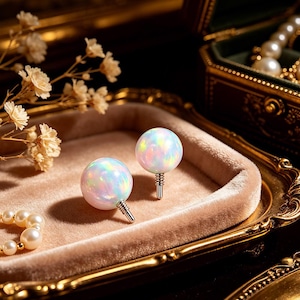 May include: Two opal sphere earrings with silver posts sit on a velvet tray. The opals show a range of colours, including pink, blue, and yellow. The earrings are displayed with other jewellery and flowers.