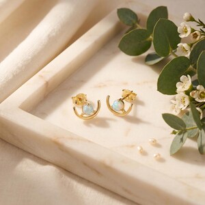 May include: Two gold-coloured earrings with a crescent moon design, each featuring a small, round, opalescent stone. The earrings are presented on a marble tray, with sprigs of greenery and small white flowers in the background.